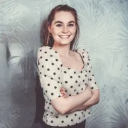 Portrait einer jungen Frau mit braunen Haaren in einem gepunkteten Oberteil, die mit geschränkten Armen vor einer Wand steht.