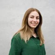 Bild einer jungen Frau vor neutralem, hellgrauem Hintergrund. Sie hat lange, glatte, mittelbraune Haare mit helleren Spitzen und trägt ein dunkelgrünes Hemd. Sie lächelt freundlich und blickt direkt in die Kamera.