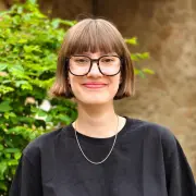 Emma Deeken vor einem grünen, braunen Hintergrund, schwarzes Shirt, Hornbrille, kurze Haare