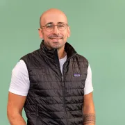 Person steht vor einem einfarbigen, hellgrünen Hintergrund. Die Person trägt eine schwarze, gesteppte ärmellose Weste mit Reißverschluss über einem weißen T‑Shirt sowie eine blaue Jeans. Beide Hände stecken in den Hosentaschen. Die Person trägt eine runde Brille und hat sehr kurz geschnittenes oder fehlendes Kopfhaar. Am rechten Unterarm ist ein geometrisches Armband‑Tattoo sichtbar. Oberkörper und Oberschenkel sind vollständig im Bild zu sehen.