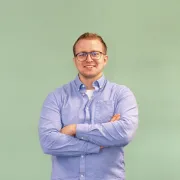 Person steht frontal vor einem einfarbig grünen Hintergrund, trägt ein hellblaues Button-down-Hemd, dunkle Stoffhose und Brille; die Arme sind vor dem Oberkörper verschränkt.