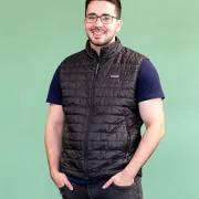 Person steht vor einem einfarbigen, hellgrünen Hintergrund. Die Person trägt eine schwarze, gesteppte ärmellose Weste mit Reißverschluss über einem dunkelblauen T‑Shirt sowie eine dunkle Jeans. Beide Hände stecken in den Hosentaschen. Die Person trägt eine eckige Brille und hat sehr kurz geschnittenes Kopfhaar. 